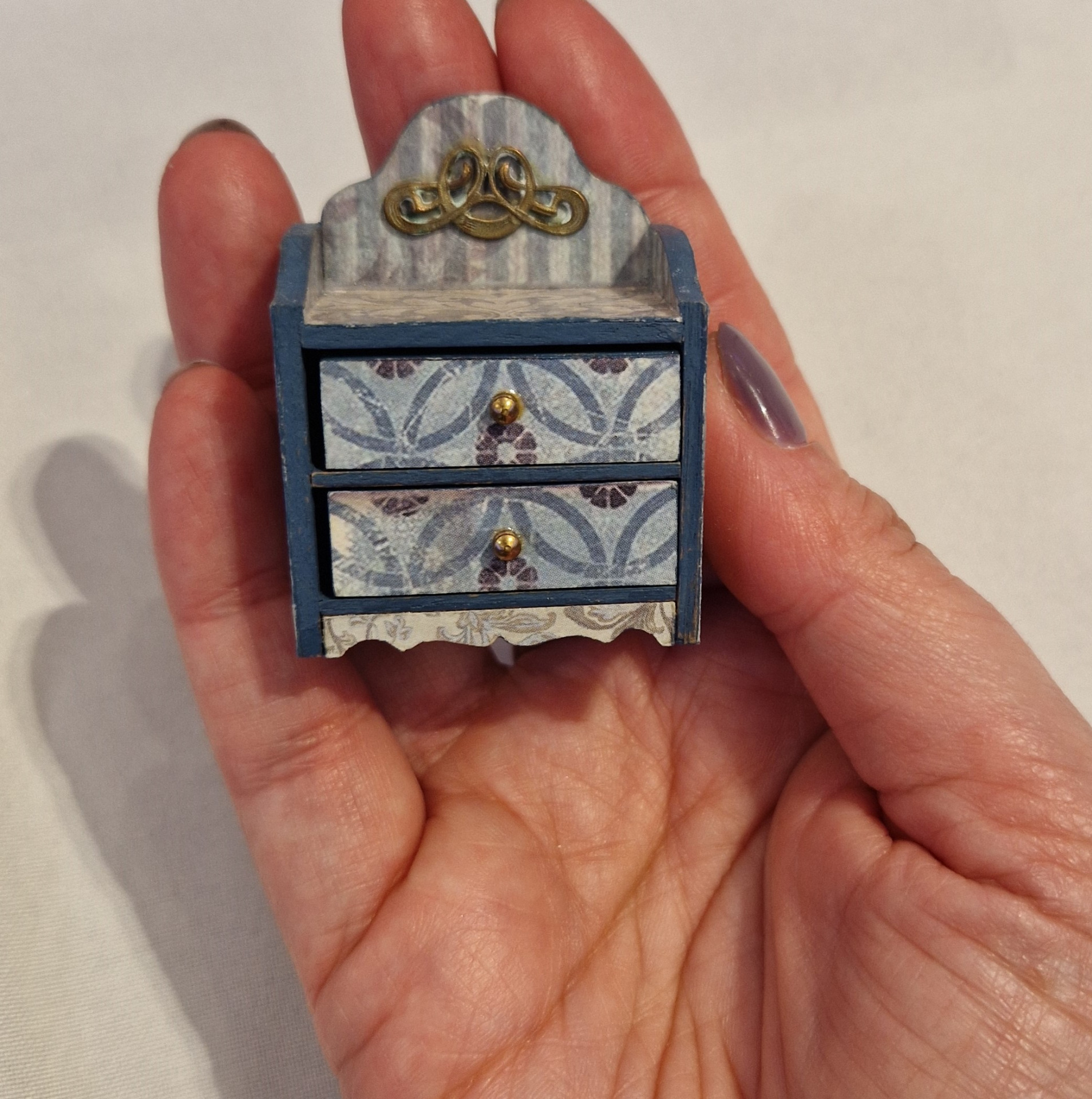 Mano sostiene una mini cómoda azul con tres cajones decorados y tiradores dorados.