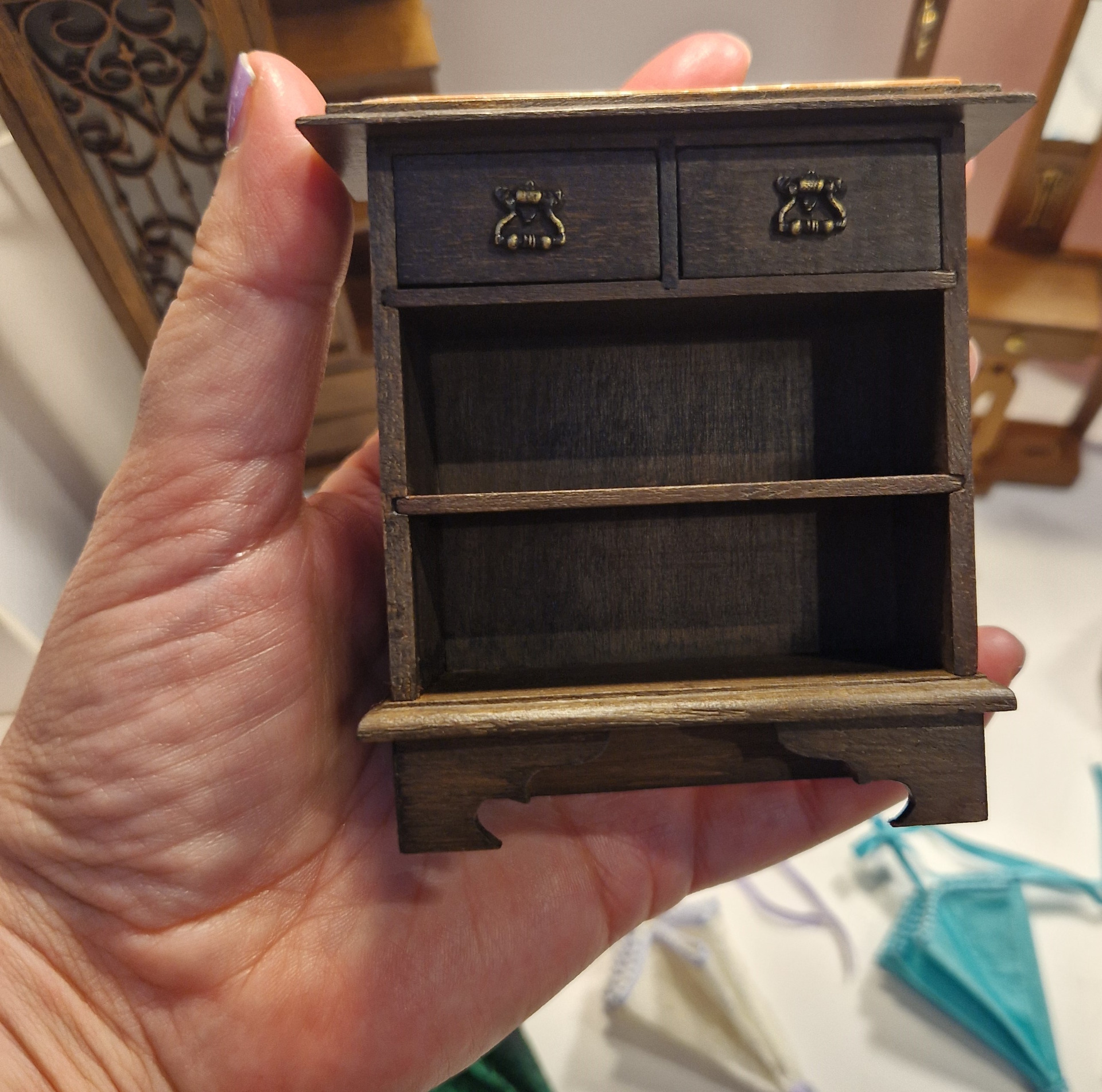 Mano sostiene una pequeña caja de madera oscura con dos cajones y herrajes decorativos metálicos.