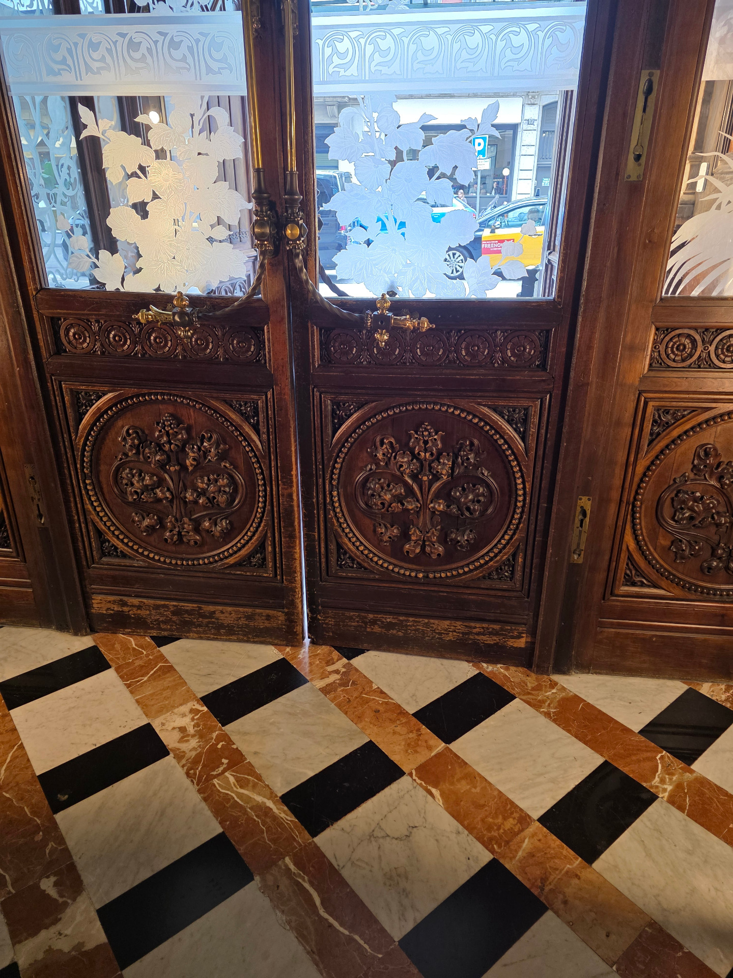 Puertas de madera tallada con paneles circulares, vidrio con flores blancas y suelo de mármol ajedrezado, entrada al Catalonia Plaça Catalunya.
