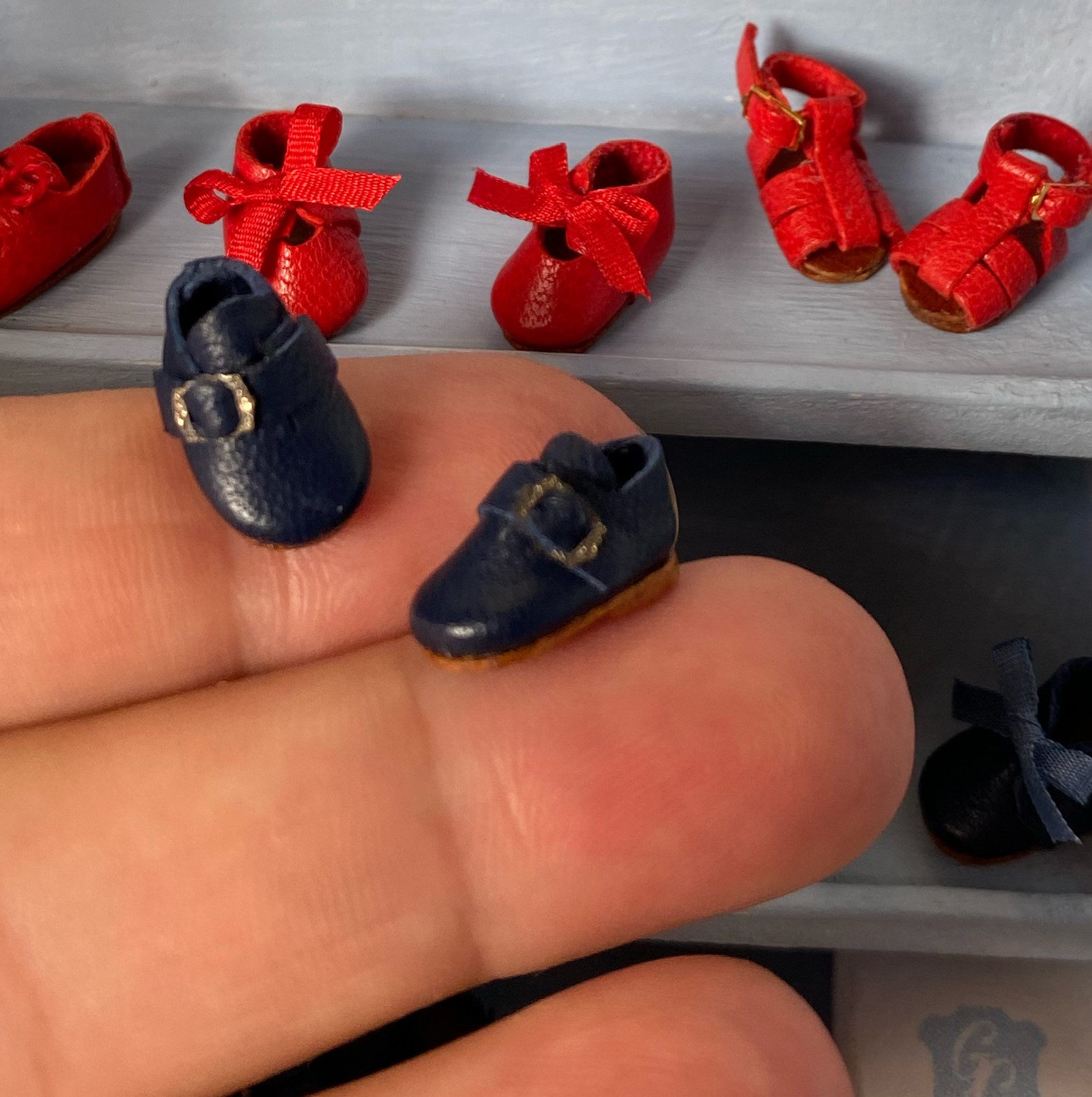 Mano sostiene pequeños zapatos negros y rojos con lazos, sobre una superficie gris clara.